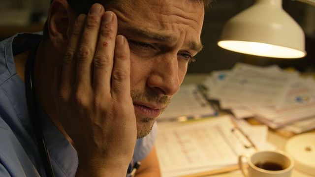 Male clinician rubbing face during night shift over stacked paperwork and coffee, fatigue