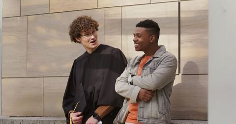 Graduation joy between diverse college friends chatting by stone wall, mortarboard in hand