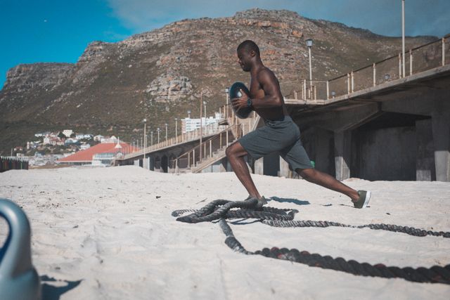 African American man engaging in a fitness routine on a sandy beach, using weights and ropes. The backdrop includes mountains and a bridge, suggesting a scenic and motivating environment for outdoor exercise. Ideal for promoting healthy lifestyles, fitness programs, outdoor workout routines, and athletic training.