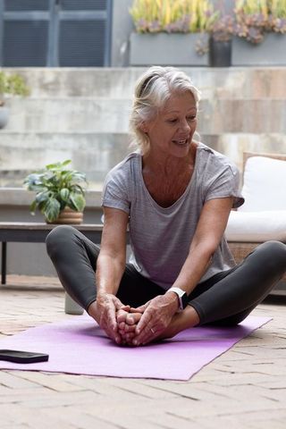 Senior woman practicing butterfly stretch on patio mat for outdoor yoga wellness wearing smartwatch