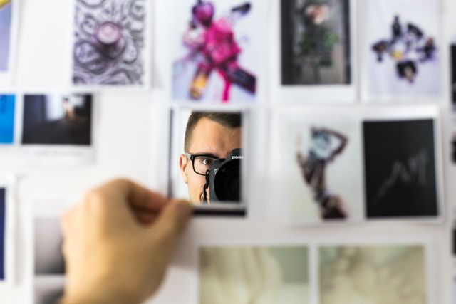 Person holding a photograph on a board covered with various pictures and artworks, reflecting on it. Ideal for use in creative projects, blogs or articles about inspiration, memory preservation, personal reflection, or photography.