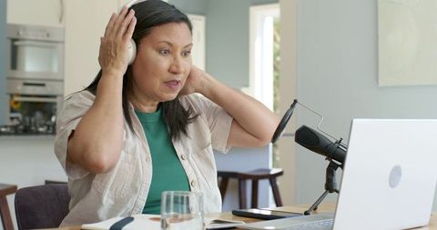 Mature asian woman recording podcast in home kitchen adjusting headphones