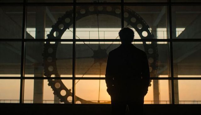 Silhouetted man standing before large industrial gear framed by paned glass at sunset
