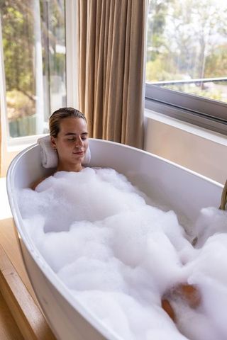 Woman relaxing in freestanding bathtub with thick foam by sunlit window in modern bathroom