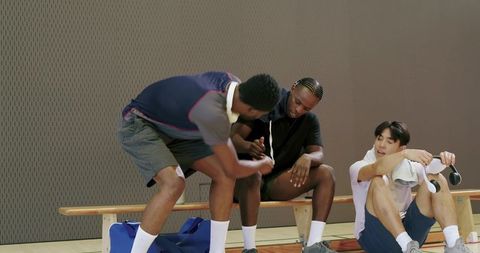 Diverse male athletes shaking hands sharing headphones team camaraderie on gym bench