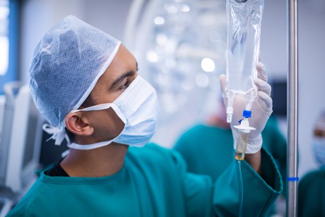 Surgeon adjusting IV drip in operating room, wearing surgical mask and scrubs. Ideal for use in healthcare, medical, and hospital-related content, illustrating surgical procedures, medical professionals at work, and patient care.