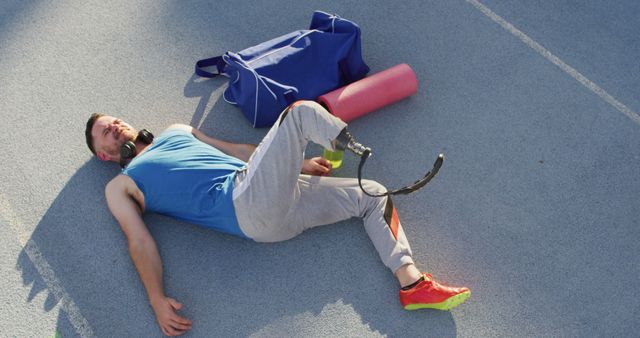 Caucasian disabled male athlete with running blade wearing headphones and stretching. professional runner training at sports stadium.