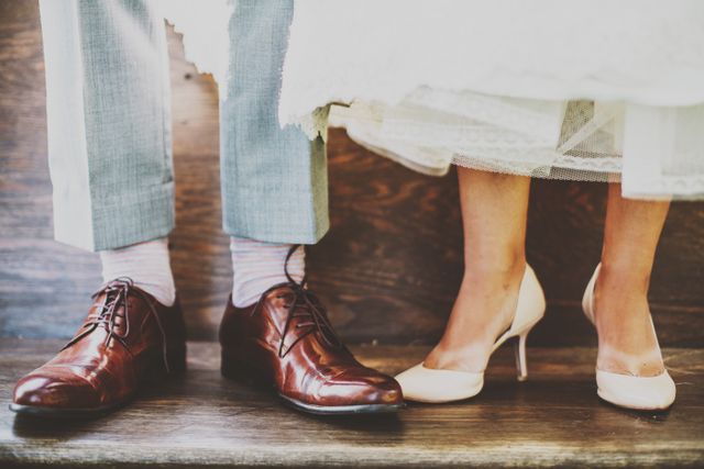 Newlyweds' feet pictured with elegant shoes on their wedding day. The groom's brown leather dress shoes complement his grey suit, while the bride's cream-colored high heels match her bridal attire. This can be used for wedding invitations, bridal magazines, fashion articles, or marriage announcement designs.