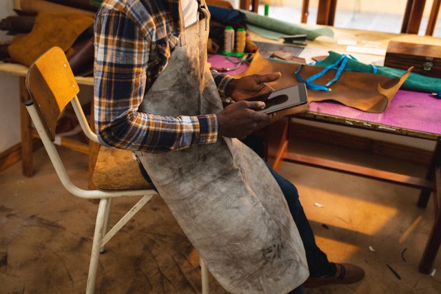 African American craftsman using digital tablet while working in a workshop. Ideal for illustrating themes of small business, craftsmanship, technology integration in traditional crafts, and creative workspaces. Useful for articles, blogs, and advertisements focusing on artisans, manual work, and the blend of technology with traditional crafts.