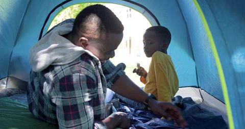 African american father and son camping in backyard tent arranging sleeping bag sharing snack