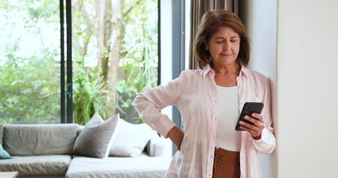 Senior woman relaxing at home using smartphone by large window