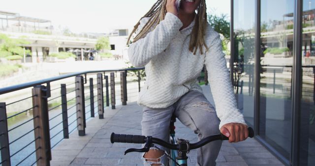 Young man with dreadlocks rides bicycle while talking on phone in urban area near glass building and rails. Suitable for content on modern lifestyle, multitasking, communication, transportation, and urban living.