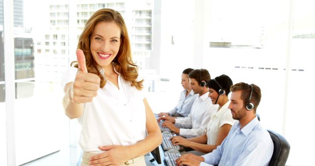 Image shows a cheerful businesswoman giving a thumbs up in a modern call center. Several colleagues with headsets are working diligently on computers in the background. Ideal for illustrating positive work environments, customer service excellence, teamwork, and professional office scenarios.