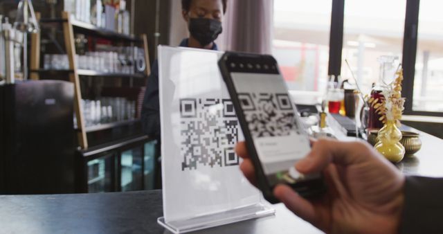 African american woman in face mask serving customer reading qr code with smartphone at cafe bar. service and technology at independent cafe business during covid19 coronavirus pandemic.