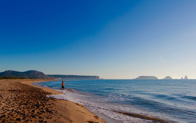 Individual strolling on quiet beach during sunrise, with clear blue sky and gentle waves. Ideal for advertisements promoting travel, vacation resorts, relaxation, or wellness retreats.