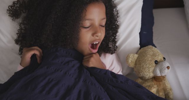 Child with curly hair yawning and pulling covers in bed while holding a teddy bear. Ideal for topics related to childhood, bedtime routines, cozy moments, and sleep habits. Perfect for use in blogs, articles or advertisements focusing on children's products, sleep aids, or parenting.