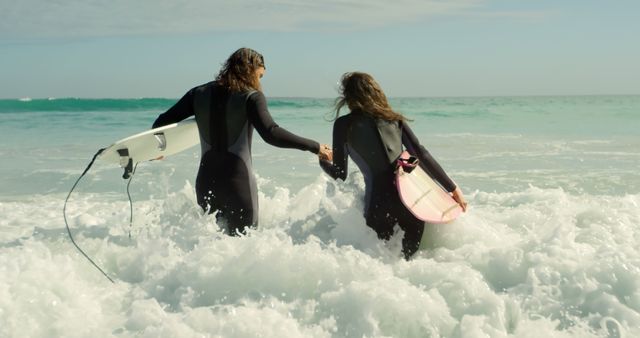 Two women in wetsuits holding surfboards walking into ocean waves, representing friendship and adventure. Ideal for content related to surfing, beach activities, water sports, and women empowerment. Useful in travel blogs, sports magazines, promotional materials, or social media posts highlighting outdoor activities.