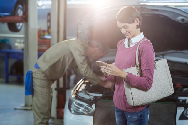 Woman standing in auto repair shop texting on her mobile phone while mechanic works on car in background. Useful for illustrating automotive services, customer satisfaction, technology use in everyday life, and vehicle maintenance.