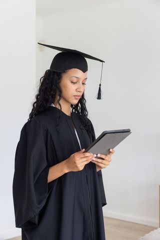 Female graduate standing in cap and gown holding tablet and reading for virtual commencement