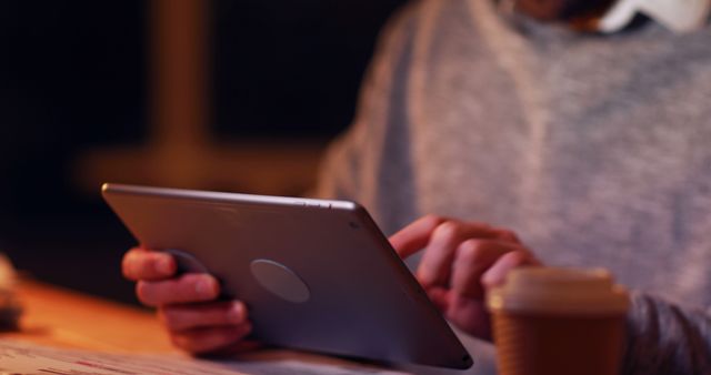 This image shows a man working on a tablet during nighttime with a cup of coffee nearby. He is focusing on the screen and wearing a casual sweater, suggesting a relaxed yet productive environment. The image is perfect for illustrating topics related to remote work, productivity, technology use, evening work, or caffeine consumption while working.