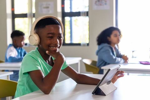 African american boy using tablet and headphones in modern classroom, smiling and focusing