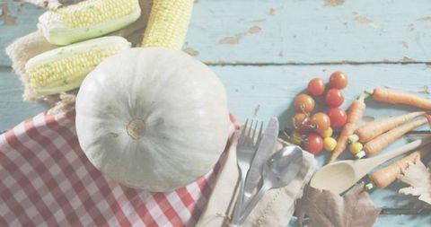 White pumpkin sitting on weathered blue table with corn, cherry tomatoes, carrots and gingham