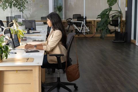 Professional woman typing at modern open-plan office desk with laptop, plants and bicycle