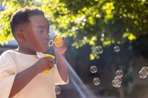African american boy blowing bubbles in sunlit backyard with yellow wand and bokeh