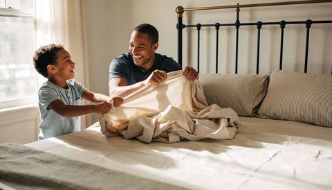 Playful father and son making bed together tugging duvet in sunlit bedroom family bonding
