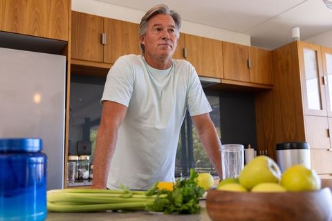 Senior man preparing green smoothie in modern kitchen with fresh pears celery herbs