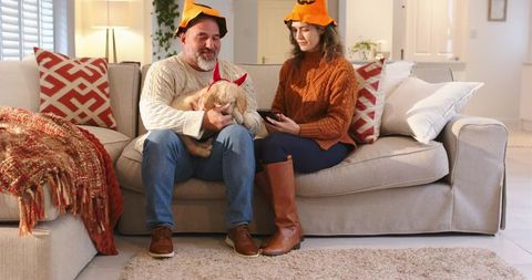 Mature couple wearing pumpkin hats sitting on couch with red-horned puppy using tablet