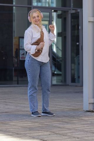 Young woman wearing pink headphones and brown crossbody bag smiling outside office building