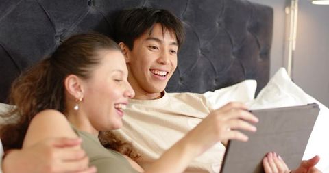 Couple relaxing on bed sharing tablet and laughing in cozy romantic bedroom