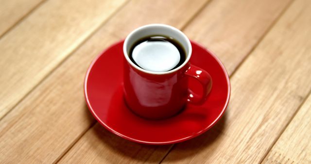 A red cup filled with coffee or tea sits on a matching saucer atop a wooden table, with copy space. Its vibrant color contrasts with the warm tones of the wood, suggesting a cozy break or morning routine.