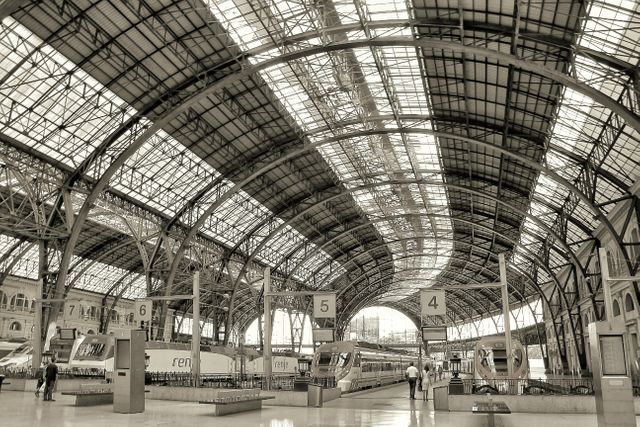 Depicts interior of a large, modern train station with grand steel and glass architecture in black and white. Train tracks and platforms are visible with some passengers in the distance. Perfect for use in travel brochures, transportation advertisements, infrastructure presentations, urban development content, and architectural portfolios.