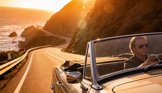 Driving convertible along winding coastal cliff road at golden hour with sunglasses driver, vintage