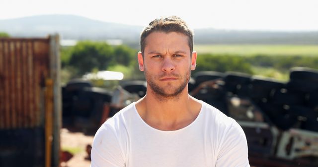 Man is standing outside in a rural area, wearig casual white t-shirt with trees and open land in the background. Photo can be used to convey themes of rural lifestyle, simplicity, or outdoor experiences.