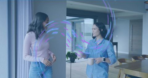 Two women chatting with holographic light streaks holding mugs in minimalist apartment