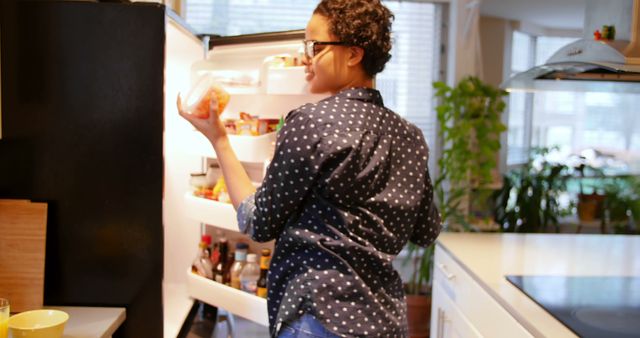 Young woman with curly hair wearing blue polka dot shirt looking inside a refrigerator in a contemporary kitchen. Scene reflects a home environment, perfect for themes of cooking, meal prep, lifestyle, or domestic tasks. Suitable for illustrating articles, websites, or advertisements related to kitchen appliances, healthy eating, grocery shopping, and everyday life.