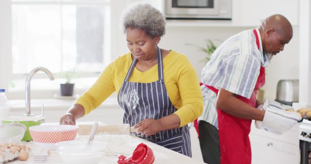 Elderly couple baking together in modern kitchen, emphasizing teamwork and home activities. Elderly man wearing red apron handling oven tray, woman focusing on dough preparation. Bright, contemporary kitchen setting with white cabinetry and natural light. Ideal for demonstrating joyful senior lifestyle, domestic bliss, and family togetherness. Useful for advertising healthy home living, senior care services, or kitchen products.