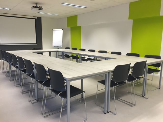 Conference room showcasing modern design with green accent walls and U-shaped table setup, surrounded by neat black chairs. Space includes a presentation screen and a flip chart, ideal for corporate meetings, presentations, and training sessions. Suitable for illustrating corporate training, business meetings, and office environments.