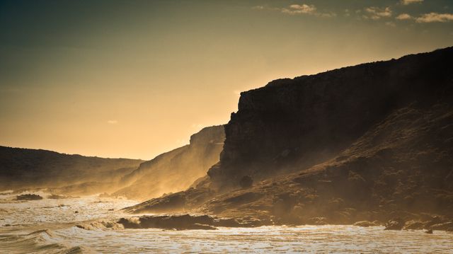Waves crashing against rugged cliffs of a coast illuminated by the warm glow of the setting sun. Silhouetted rocks and golden mist add to the dramatic beauty of the scene. Ideal for nature-themed projects, travel promotions, inspirational posters, or backgrounds for presentations highlighting natural scenery.