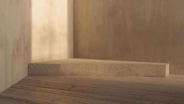 Warm low-angle sunlight on minimal plinth and textured plaster corner with wood floor