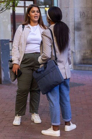Female friends chatting on city sidewalk carrying shoulder bags and smartphone