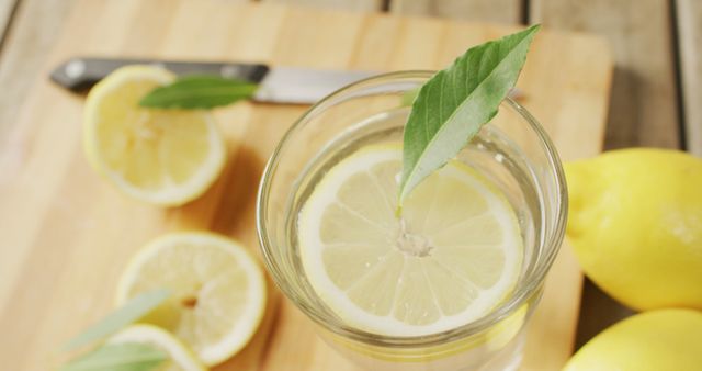 Refreshing lemon water garnished with fresh leaves in glass on wooden table. Bright, healthful drink with whole and sliced lemons around. Ideal for content promoting healthy eating and lifestyle, beverage recipes, and summer refreshment ideas.