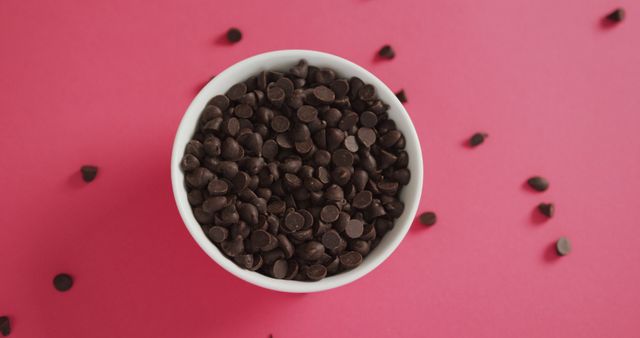 Close-up of dark chocolate chips in a white bowl on a pink background, showing details of the chocolate pieces. Perfect for use in food blogs, recipe websites, baking tutorials, or any culinary-related content. Ideal for emphasizing sweetness or dessert preparation themes.