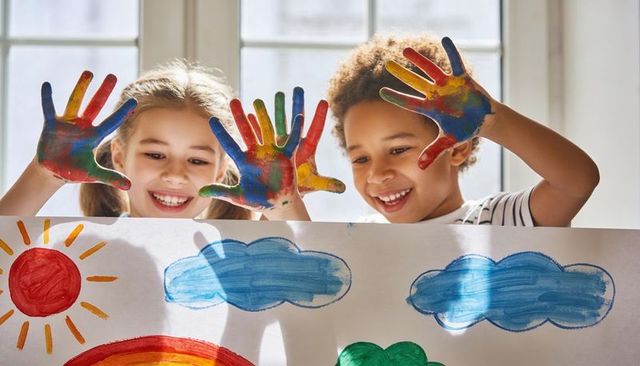 Joyful children holding colorful painted hands above rainbow artwork in bright classroom