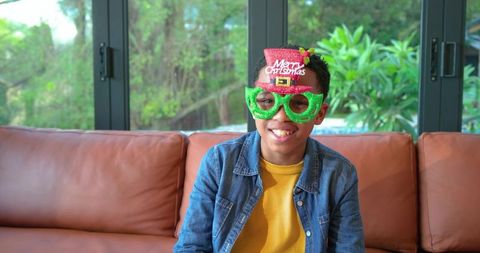 Smiling african american boy wearing glitter merry christmas glasses on leather couch holiday cheer