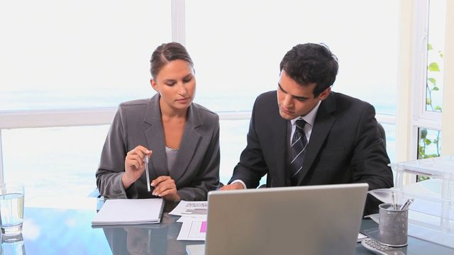 Business professionals shown intensely discussing and studying statistics in a modern office. The clear glass walls and well-lit environment create a professional corporate atmosphere. Useful for illustrating themes related to teamwork, office life, corporate meetings, work collaboration, and business analysis.