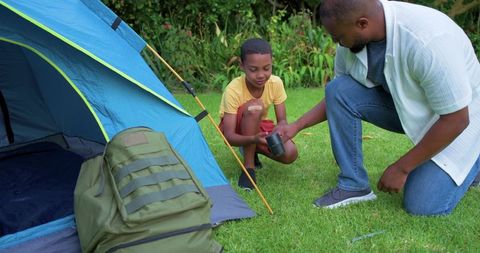 African american father and son preparing backyard camping, exchanging cup by blue tent